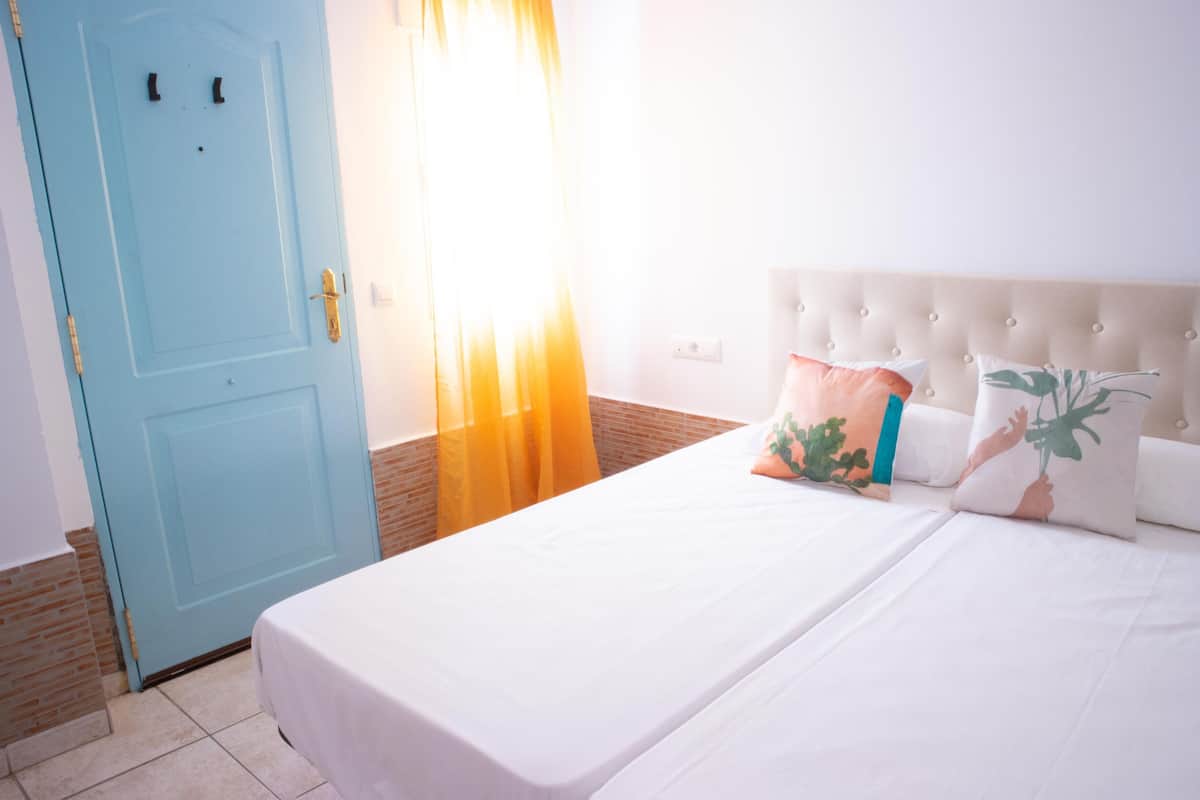 Una habitación de hotel con una puerta azul claro, una cama con ropa blanca y un cabecero capitonado.