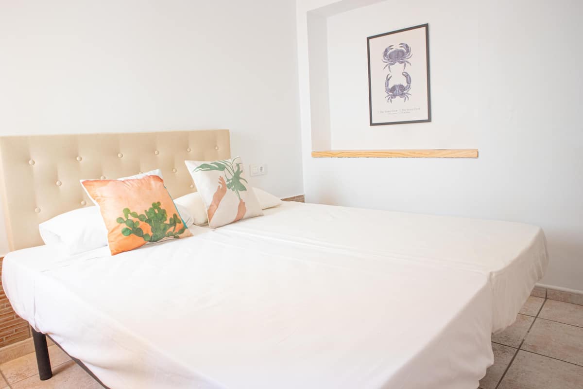 Una cama bien tendida con una colcha blanca y dos almohadones decorativos, una obra de arte enmarcada en la pared, y un estante de madera debajo.