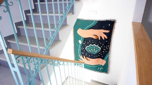 A staircase with teal railings and a wall-mounted tapestry depicting hands reaching towards a celestial scene. - Hostal Salitre (Conil de la Frontera)