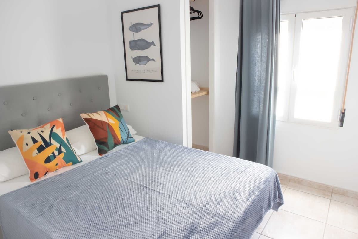 Un dormitorio con cama, almohadas, un cuadro enmarcado, una ventana con cortinas y una puerta.