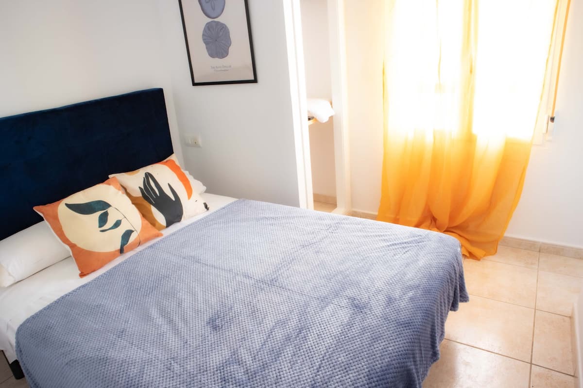Un dormitorio con cama, almohadas, cabecera, un cuadro enmarcado en la pared y ventana con cortinas.