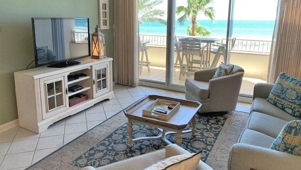 Ein Wohnzimmer mit Fernseher, Schrank, Couchtisch und Sofa. Es gibt einen Balkon mit Blick auf den Ozean und Palmen.