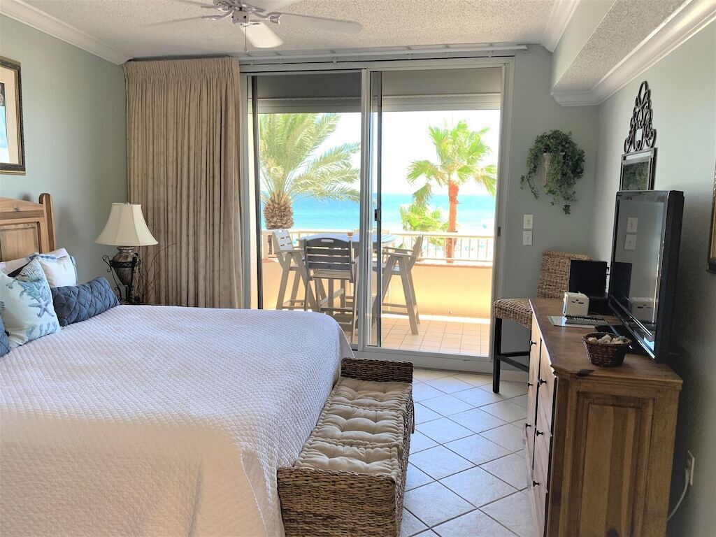A bedroom with a bed, a desk, a television, and a balcony with a view of the ocean.