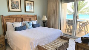 Ein Schlafzimmer mit einem großen Bett, einer Korbsitzbank, einem Nachttisch, einer Lampe und Blick auf den Strand durch eine Schiebetür.