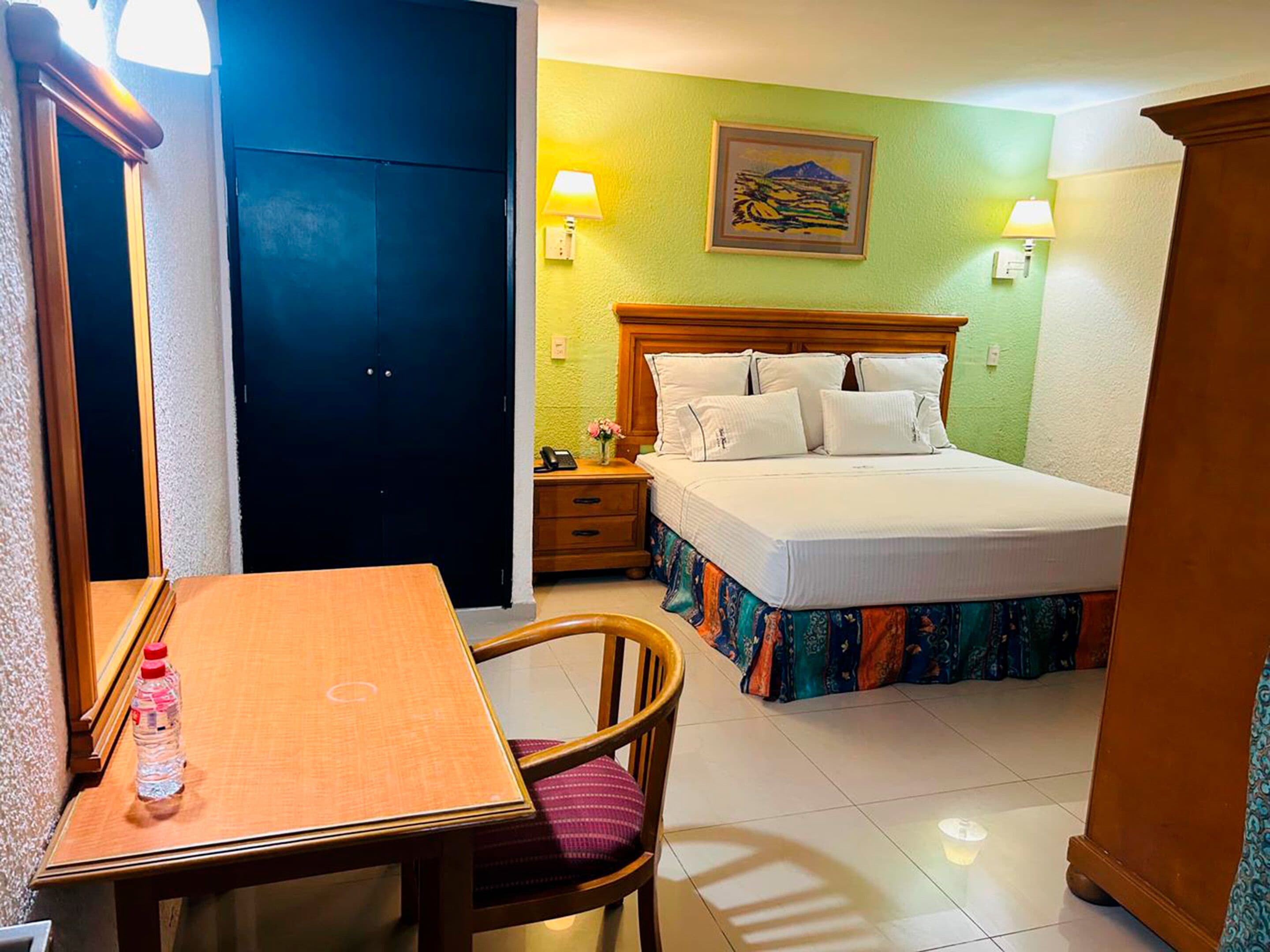 A hotel room with a bed, a desk, a chair, a nightstand, and a wall with a painting.