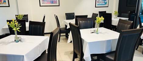 Uma sala de jantar com mesas cobertas por toalhas brancas, cadeiras pretas e vasos com flores amarelas.