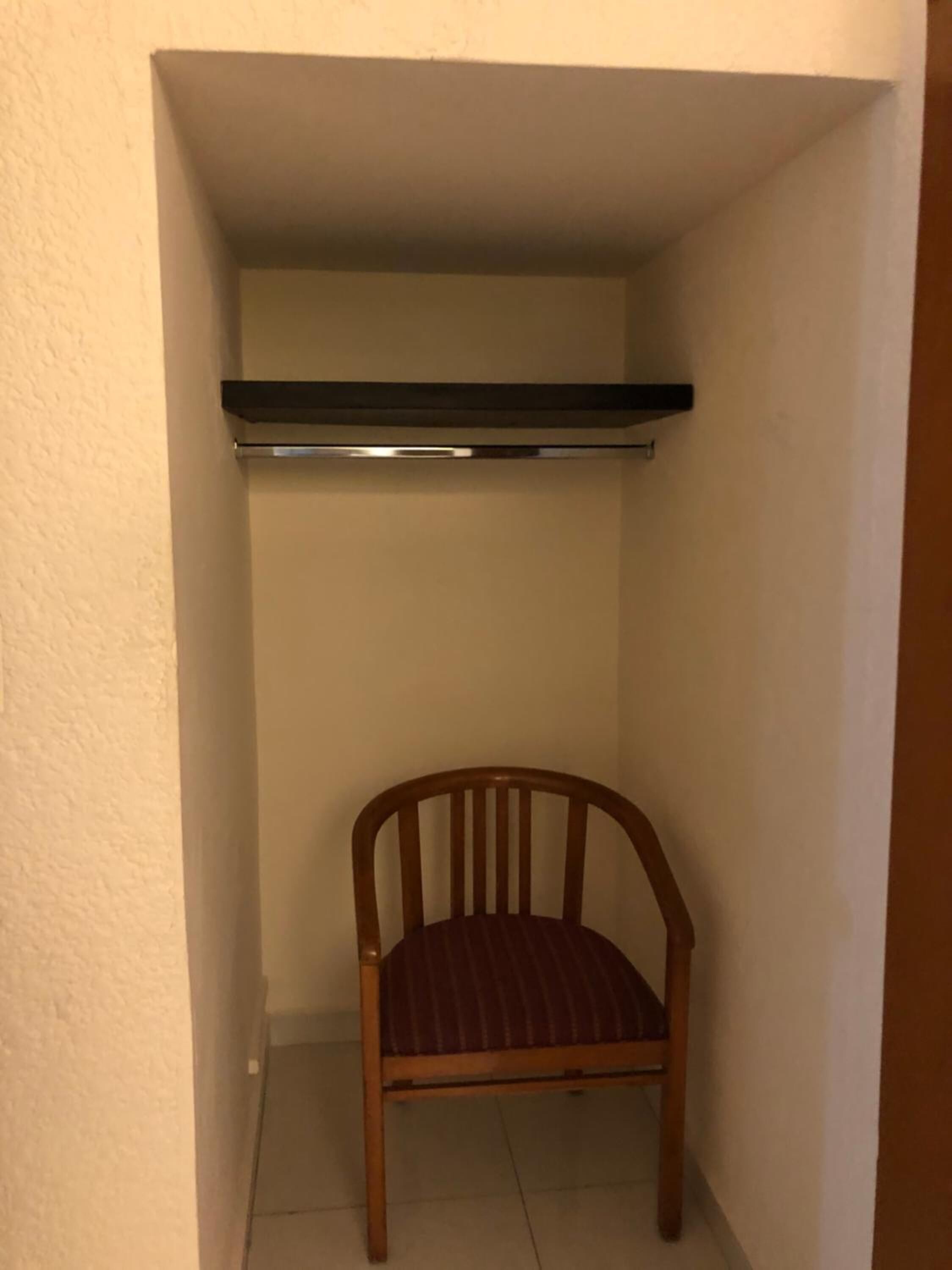 Um quarto pequeno e estreito, com uma cadeira de madeira e uma prateleira preta.