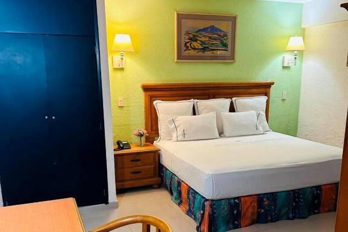 Una habitación de hotel con cama, mesita de noche, silla y un cuadro en la pared.
