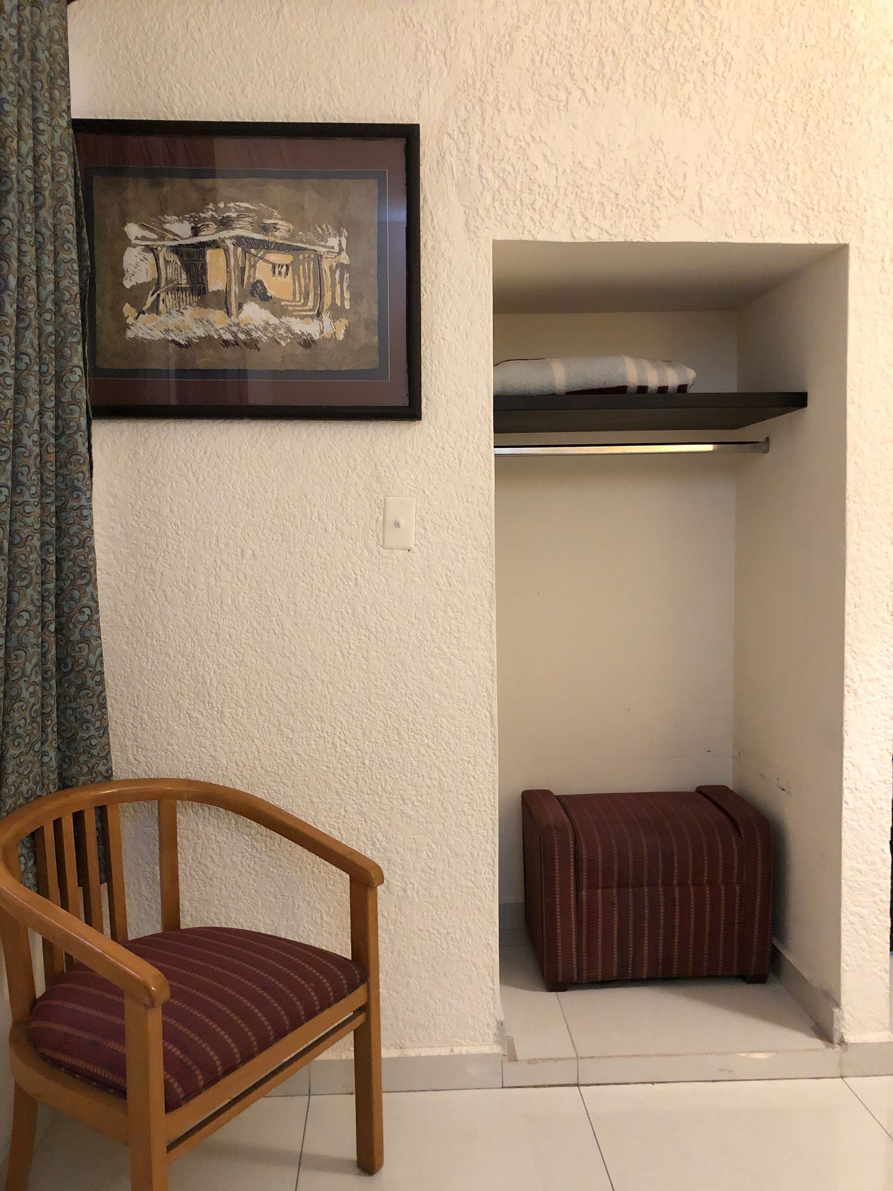 A corner room with a wooden chair, a framed picture on the wall, a small bench, and a shelf with a towel.