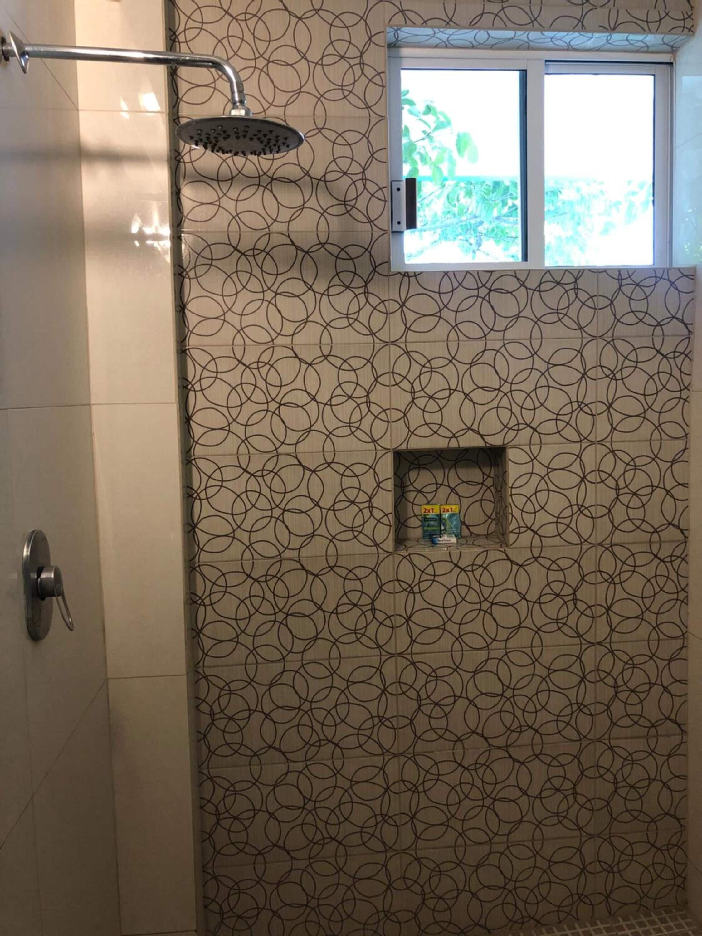 A shower with a circular pattern on the wall, a window with a view of greenery, and a shelf with toiletries.