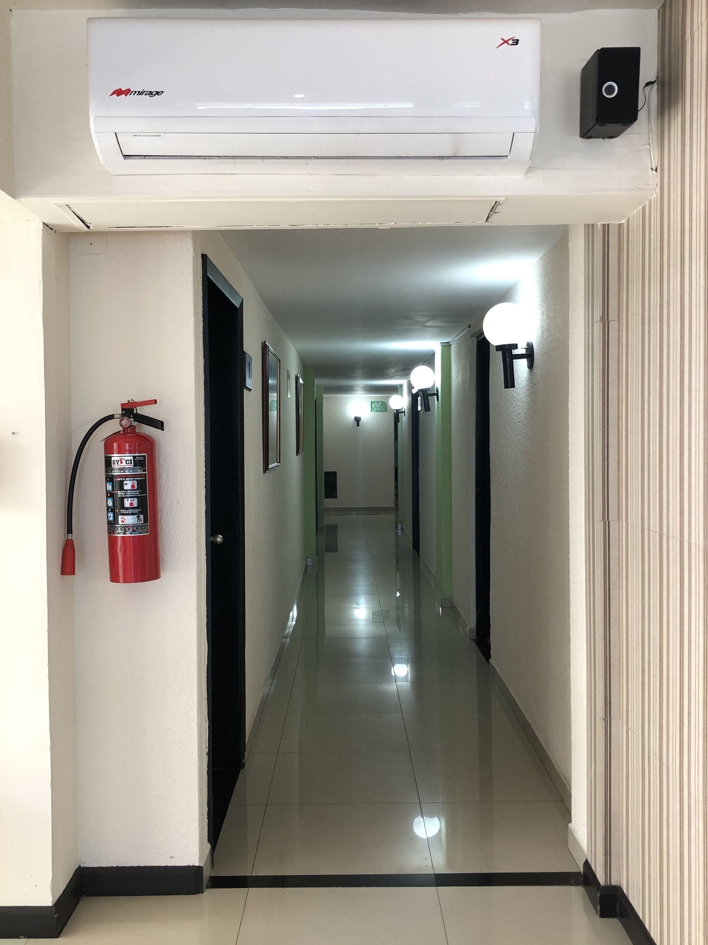 A hallway with a fire extinguisher, a ceiling-mounted air conditioning unit, and wall-mounted lights.