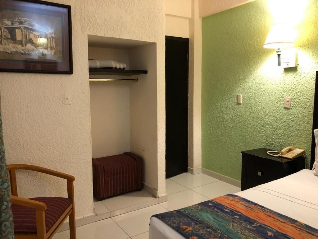 A hotel room with a bed, a chair, a small table with a phone, a shelf with a towel, and a green wall.