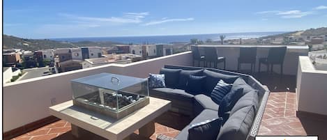 Una terraza en la azotea con una mesa de vidrio, un sofá gris y vistas al mar y las colinas.