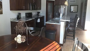 Una cocina moderna con armarios de madera oscura, electrodomésticos de acero inoxidable y encimera de mármol. Hay una mesa de comedor con sillas y un elegante soporte de metal.