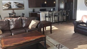 Una sala moderna con un sofá de cuero marrón oscuro, una mesa de centro de madera y una cocina con armarios oscuros y un refrigerador.