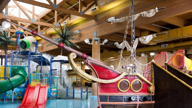Parque acuático interior con área de juegos temática de piratas, que incluye toboganes y una piscina con forma de barco.