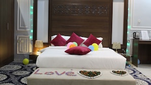A neatly made bed with a white comforter and multiple pillows, a bedside table with a lamp, and a decorative wall panel above the bed. - Laylati Al Yarmouk (Riyadh)