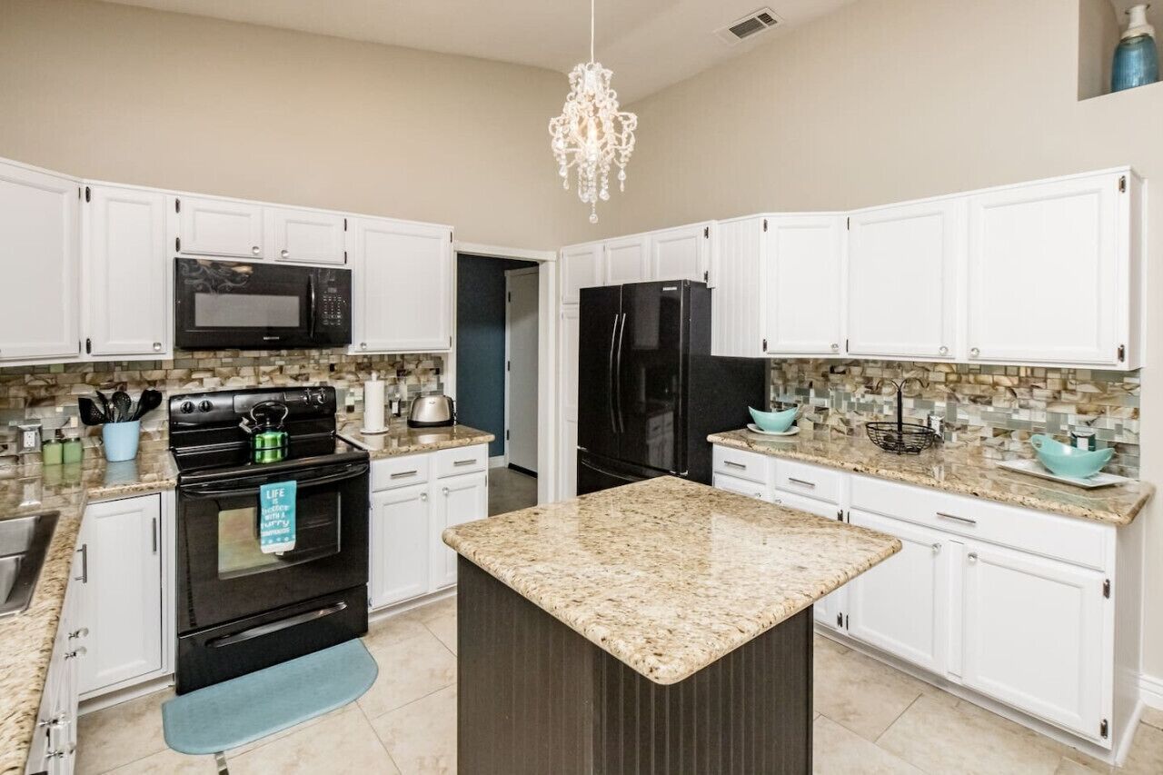 A modern kitchen with white cabinets, a granite countertop island, a black stove, a black refrigerator, and a chandelier.