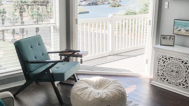 A living room with a view of a marina, a white chair, a round table, and a white pouf. - WATER VIEW! POOL! HOT TUB! SAUNA! GYM! WALK TO BEACH! PET FRIENDLY! (Corpus Christi)