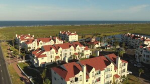 A coastal residential area with multi-story buildings, a road, and a body of water in the background. - WATER VIEW! POOL! HOT TUB! SAUNA! GYM! WALK TO BEACH! PET FRIENDLY! (Corpus Christi)