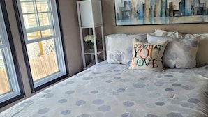 A bedroom with a large bed, a cityscape painting, and a window with a view of a wooden deck.