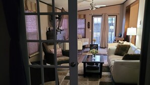 A living room with a white sofa, a coffee table, and a TV. There is a door leading to another room.