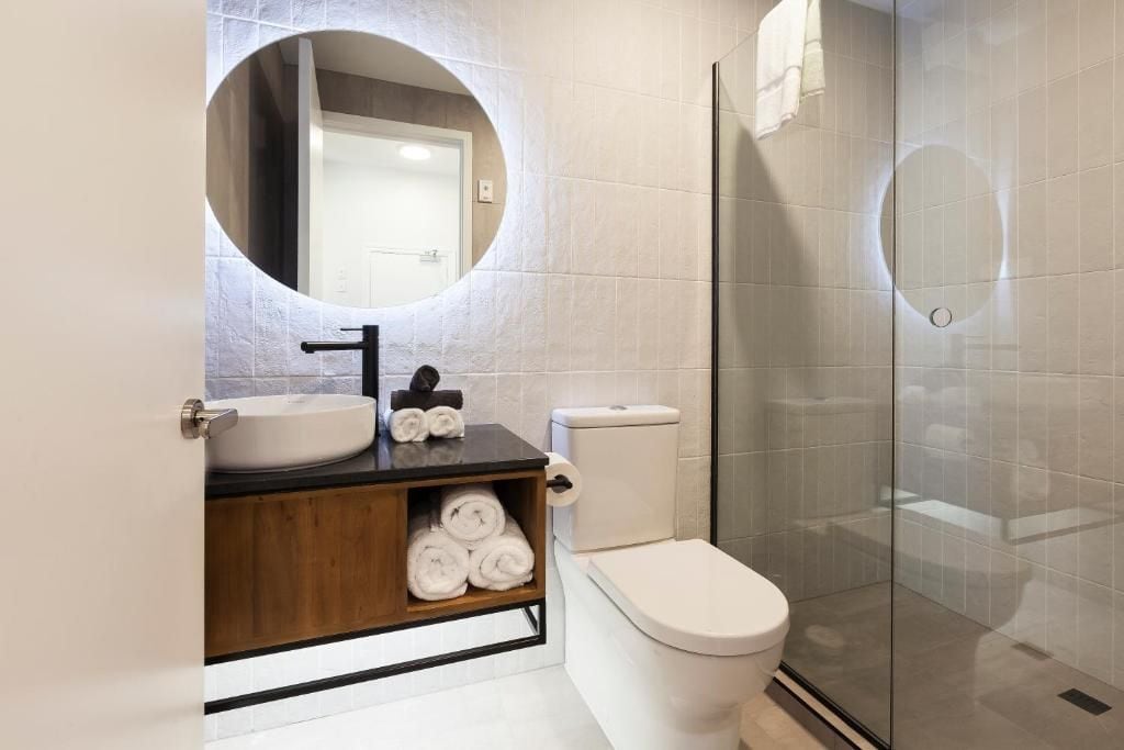 Ein modernes Badezimmer mit rundem Spiegel, Waschbecken, Toilette und Duschkabine aus Glas.
