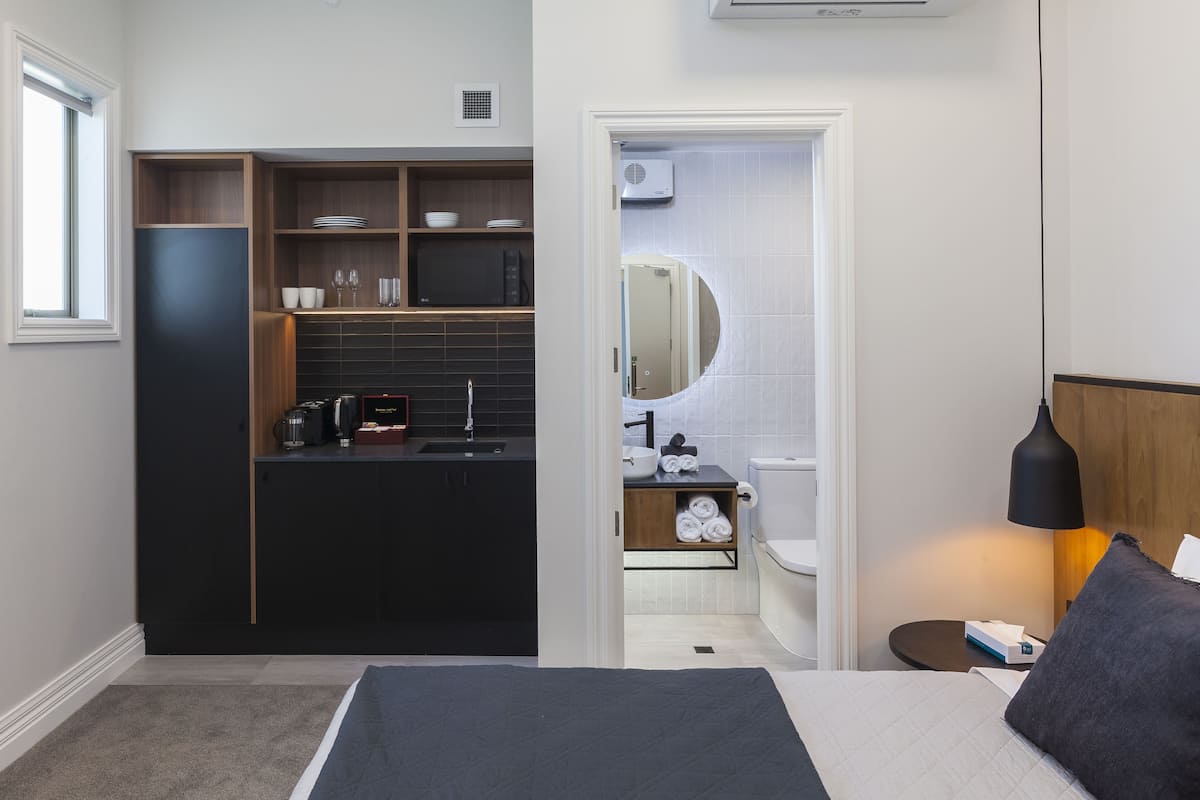 A modern hotel room with a bed, a small bedside table, a nightstand, a desk with a chair, a built-in kitchen with a sink and cabinets, a toilet, and a mirror.