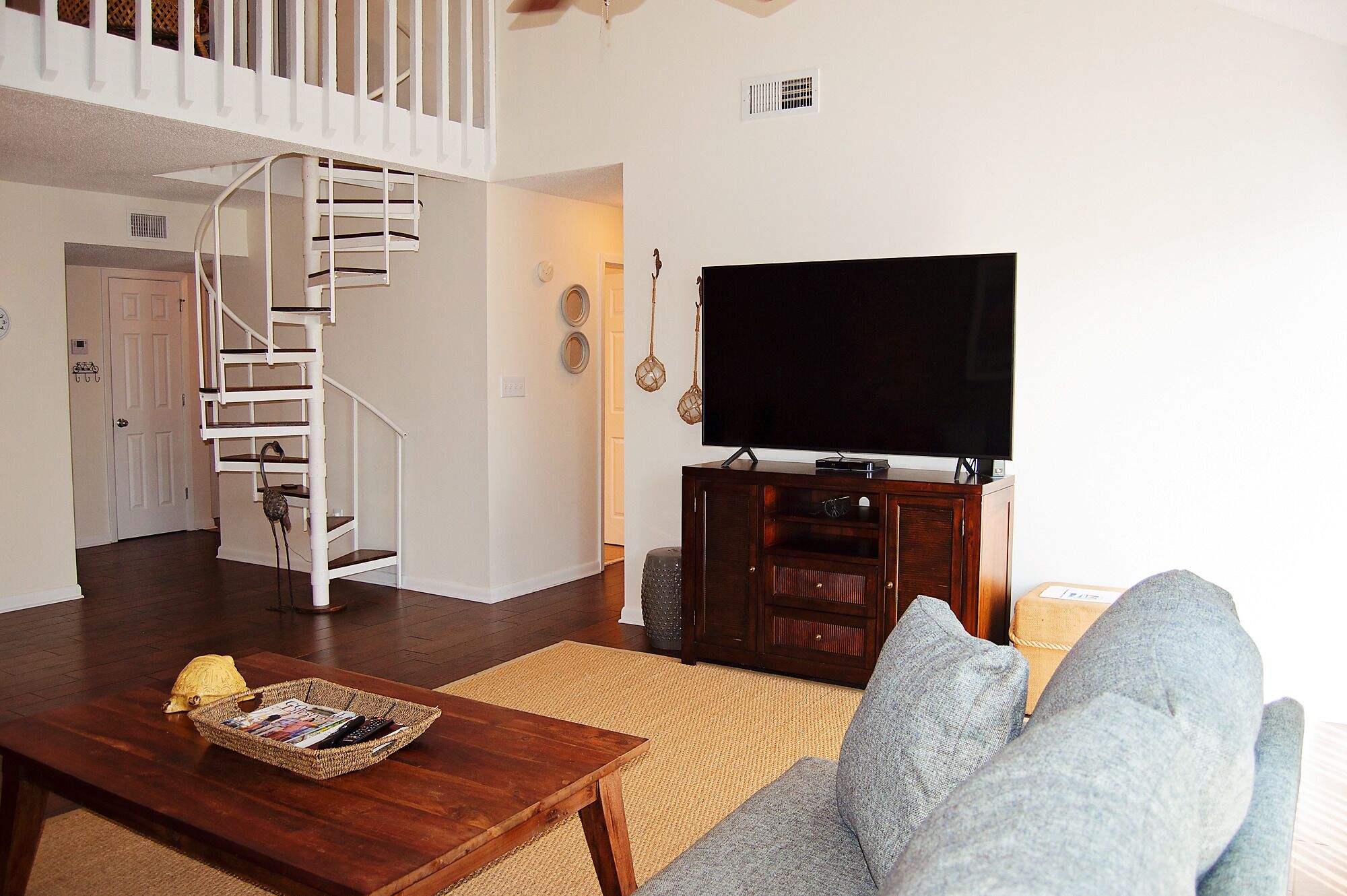Ein Wohnzimmer mit Treppe, einem Flachbildfernseher auf einem Holzboden, einem Couchtisch mit Zeitschriften und einem grauen Sofa.