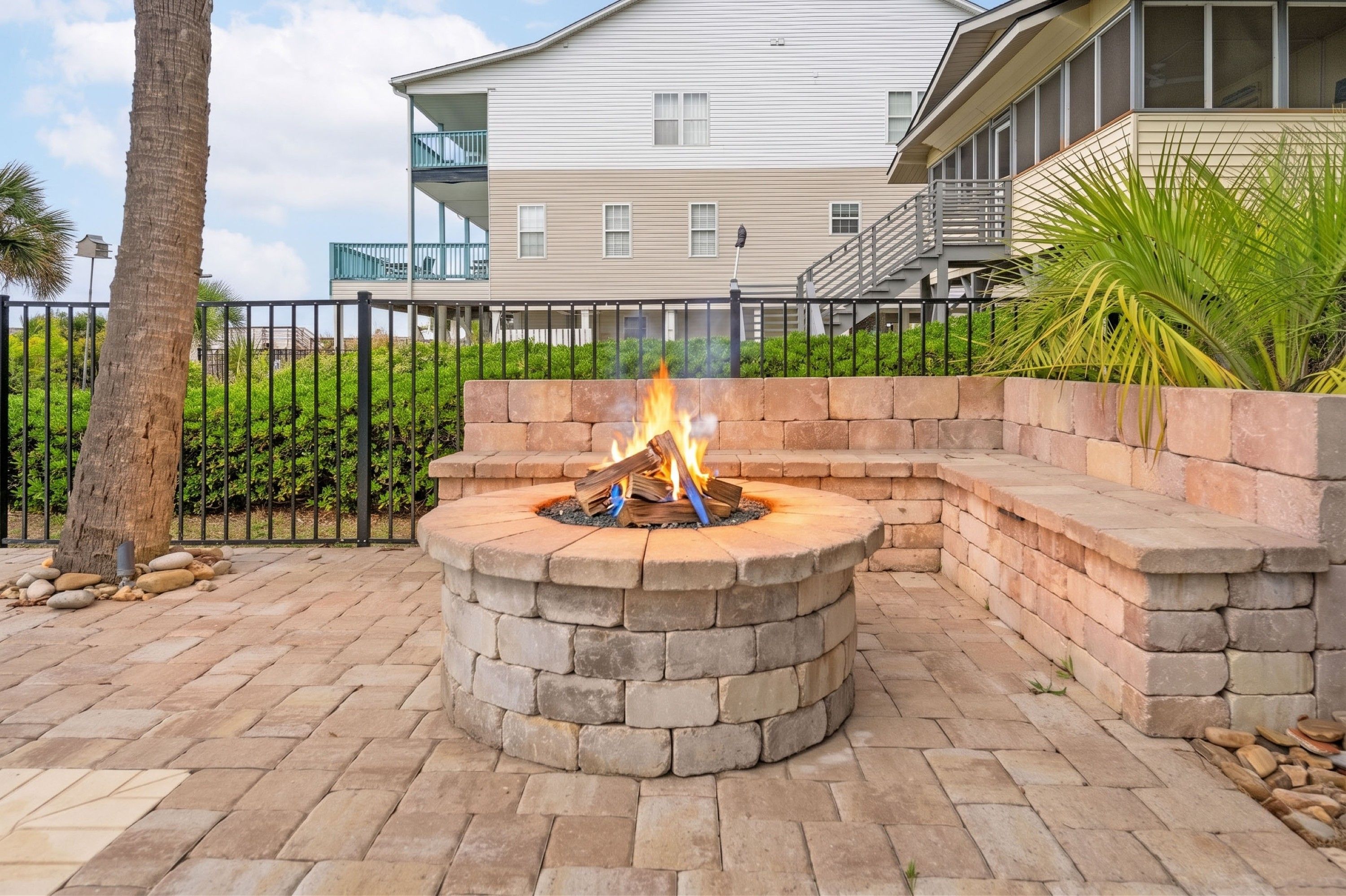 A fire pit with a stone fire surround, a metal fence, and a residential building in the background.