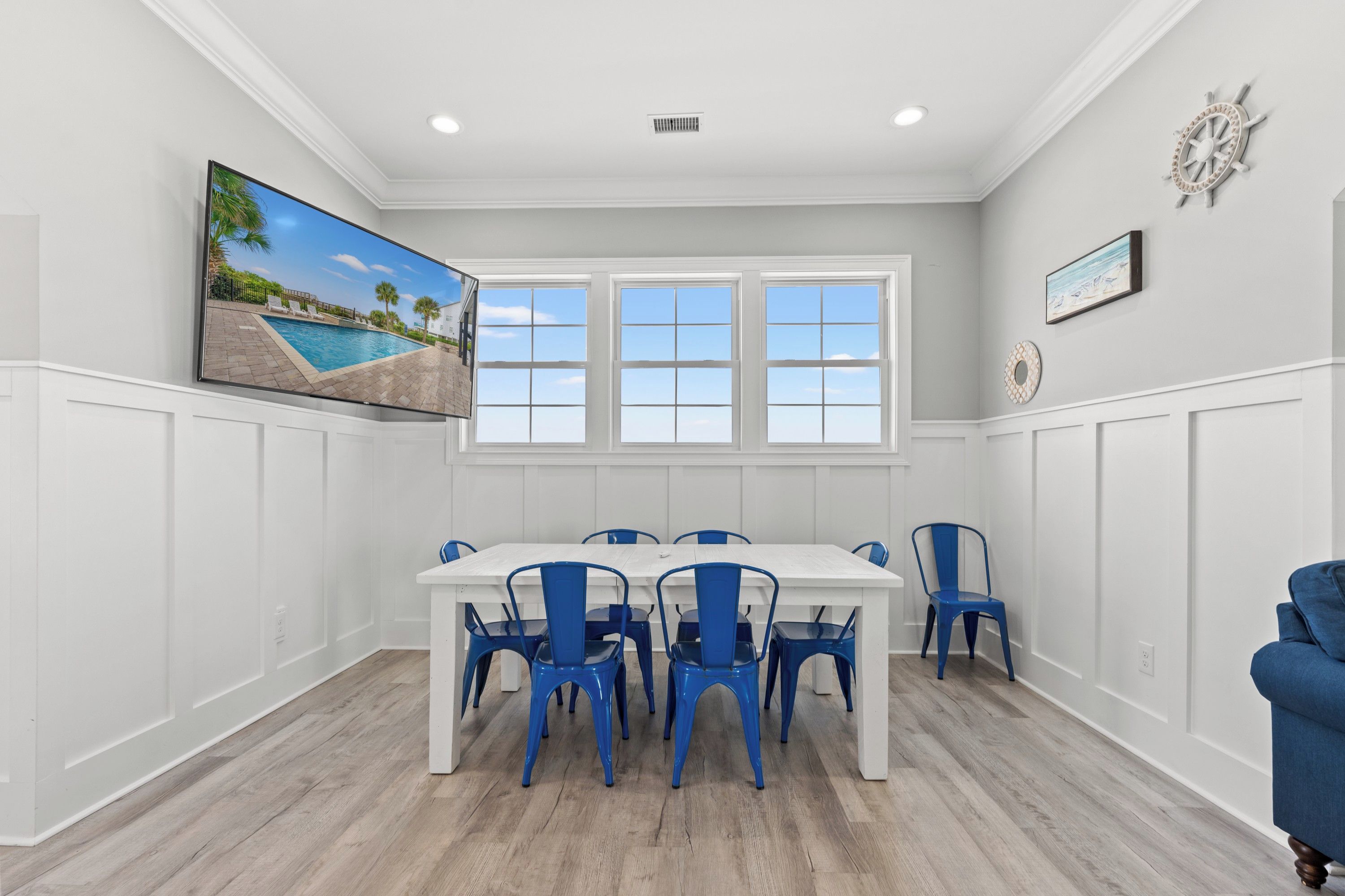 A dining room with a white table and blue chairs, large windows, and a painting of a beach scene.