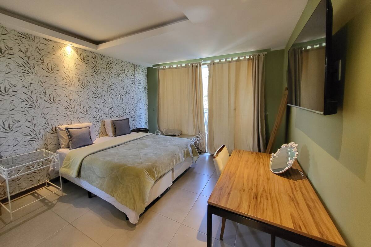 Habitación de hotel con una cama, una mesa de madera, un televisor montado en la pared y un papel tapiz estampado.