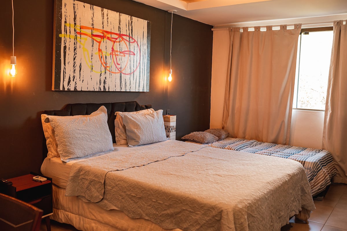Una cama doble con una colcha blanca, dos mesitas de noche, una mesita auxiliar, un cuadro abstracto en la pared y una ventana con cortinas.