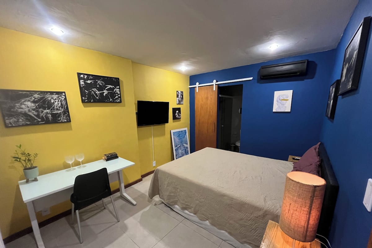 Un dormitorio con cama, escritorio, silla, planta en maceta y televisión.