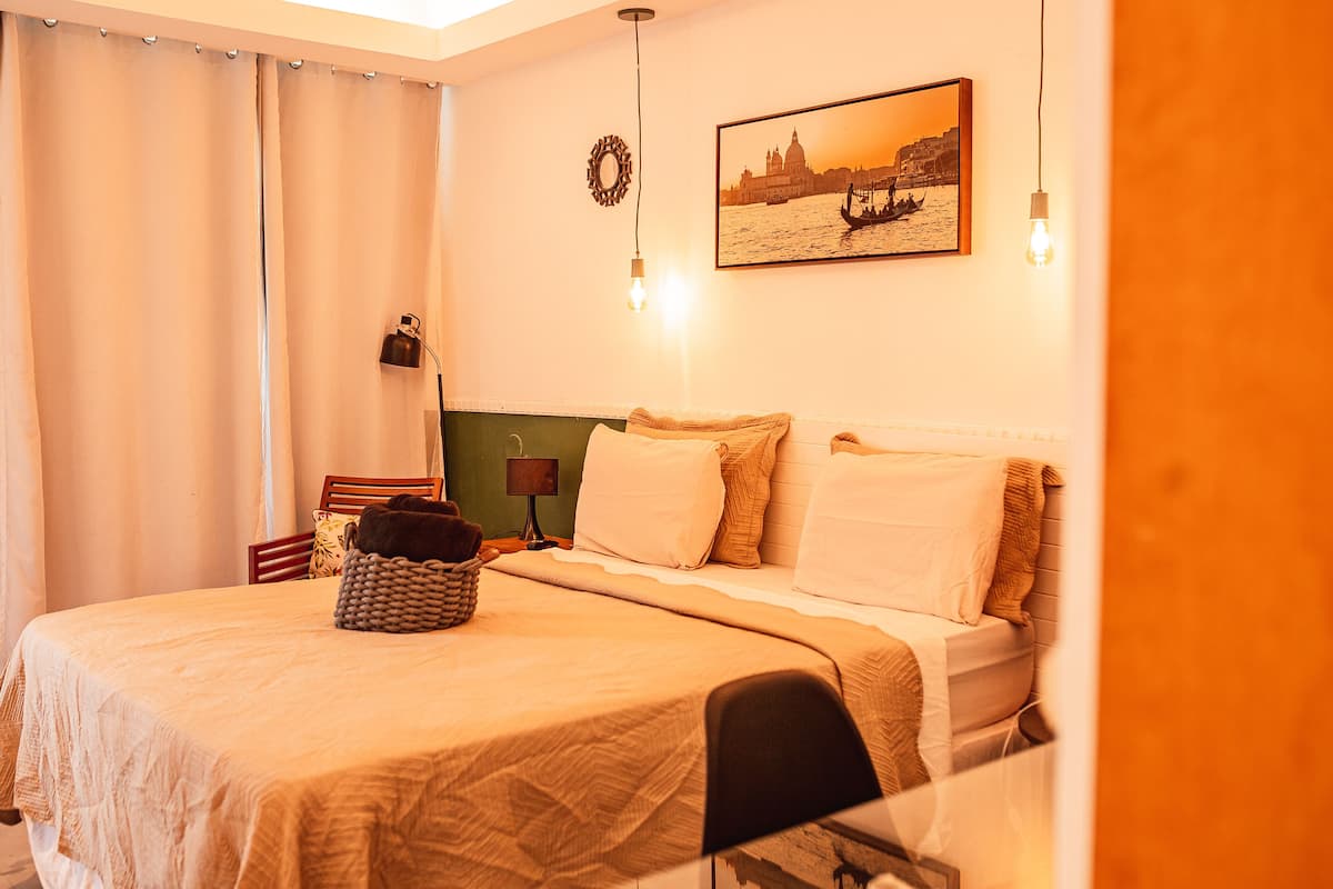 Una cama bien hecha con edredón color crema, una cesta tejida y una lámpara sobre una mesita de noche.