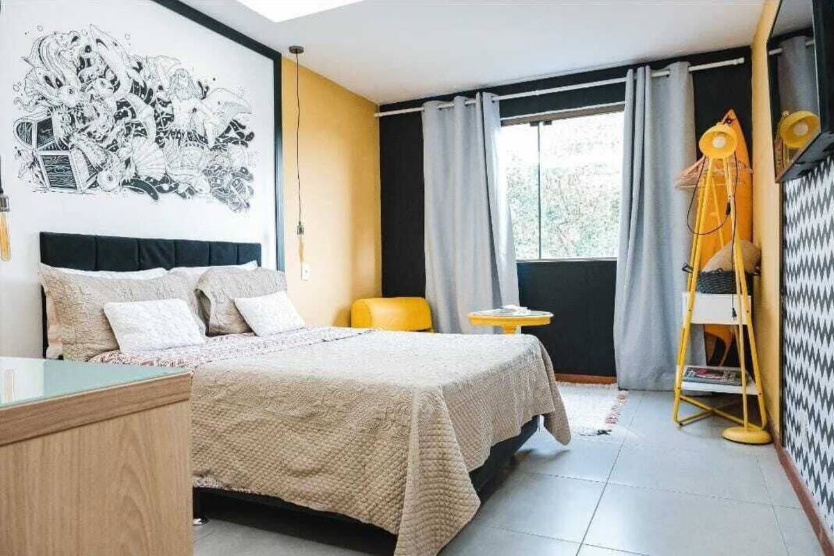 Un dormitorio con cama, una mesita, una silla, una ventana con cortinas y un cuadro blanco y negro en la pared.