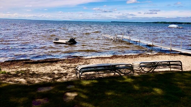 A beach with a wooden pier extending into calm waters, a small boat anchored near the pier, and two metal benches on the sandy shore. - New Listing! Lake front cottage with 100' of private sandy beach (Prudenville)