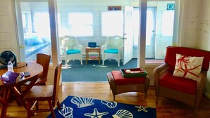 A living room with a wooden floor, a blue rug with sea shells, two white wicker chairs, a red armchair, and a small table with a laptop. - New Listing! Lake front cottage with 100' of private sandy beach (Prudenville)