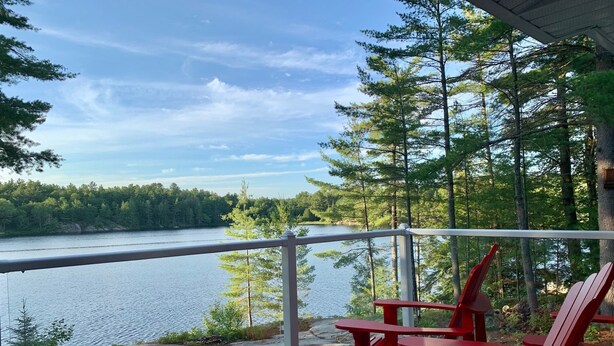 A wooden deck with red Adirondack chairs overlooking a lake surrounded by trees.