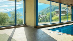 A modern indoor swimming pool with large windows offering a view of mountains and houses. - Arnica 16 by Interhome (Nendaz)