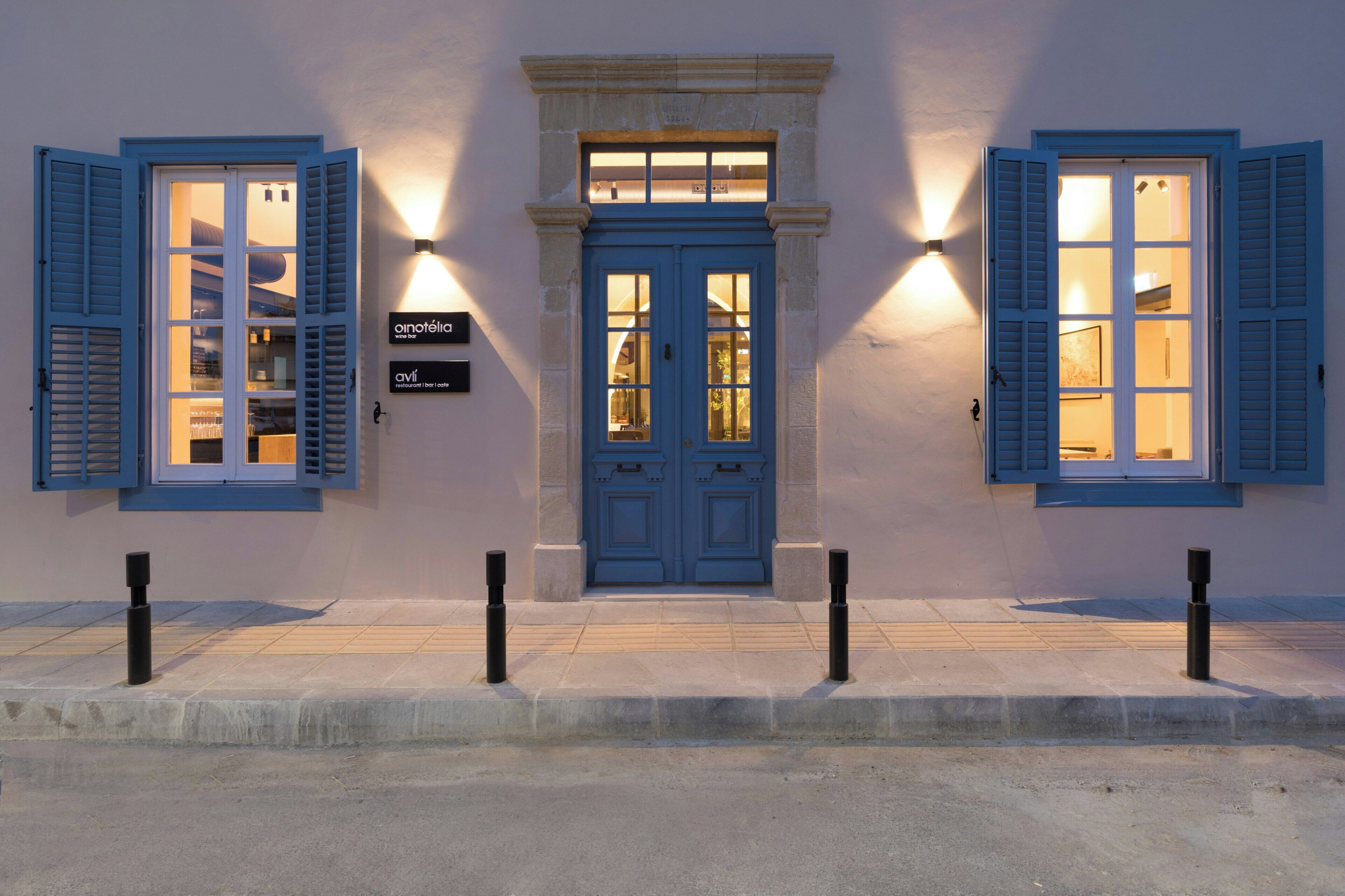 A building with a blue door and shutters, a small window above the door, and two wall-mounted lights.