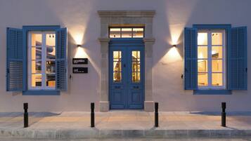 A building with a blue door and shutters, a small window above the door, and two wall-mounted lights.