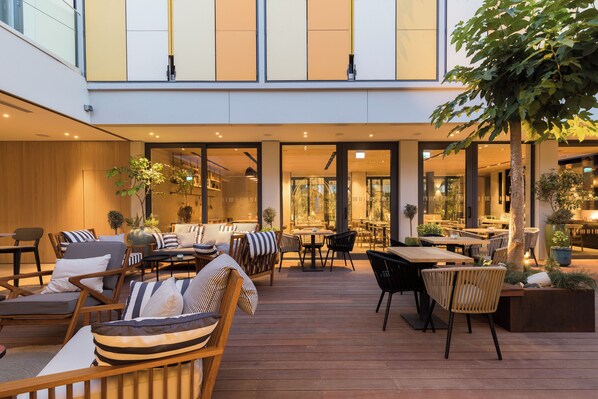 An outdoor seating area with wooden furniture and tables, surrounded by plants and trees. - Indigo Hotel - Adults Only (Larnaca)