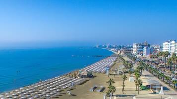 A coastal cityscape with a beach lined with umbrellas and palm trees, buildings along the shore, and a clear blue sky.