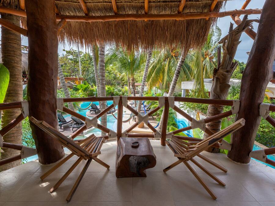 Una terraza de madera con techo de paja que da a una zona de piscina, rodeada de palmeras y sillones de descanso.