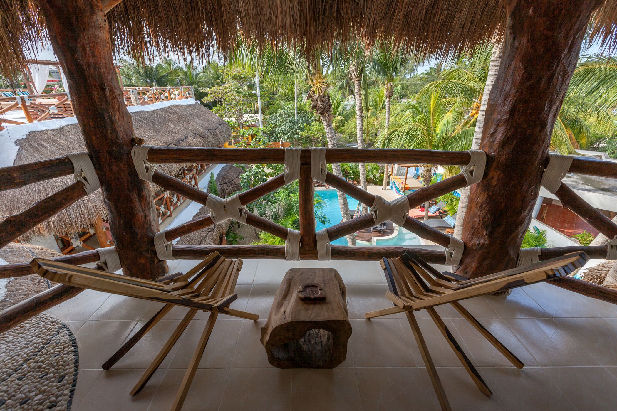 Silla hamaca de madera montada sobre un tronco central, con vista a una piscina y abundante vegetación.