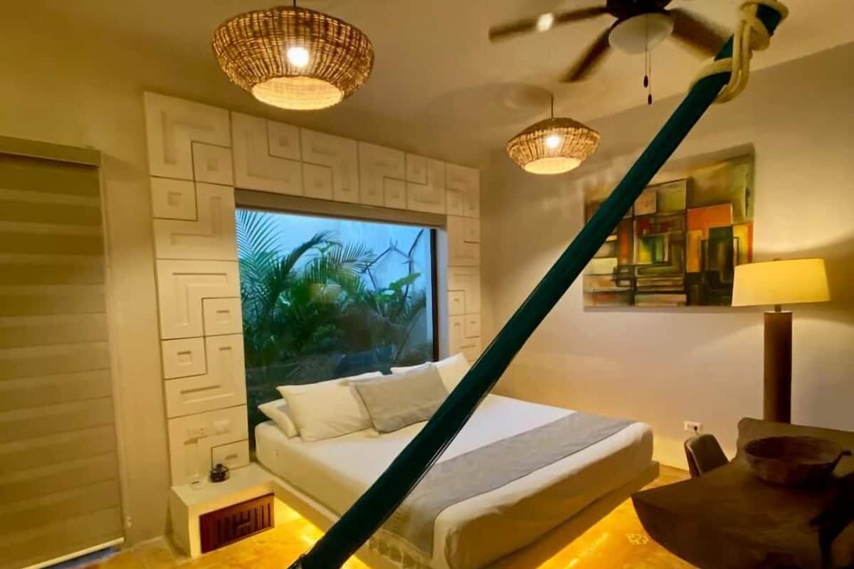 A modern bedroom with a bed, a hammock, a ceiling fan, and a large window with a view of palm trees.