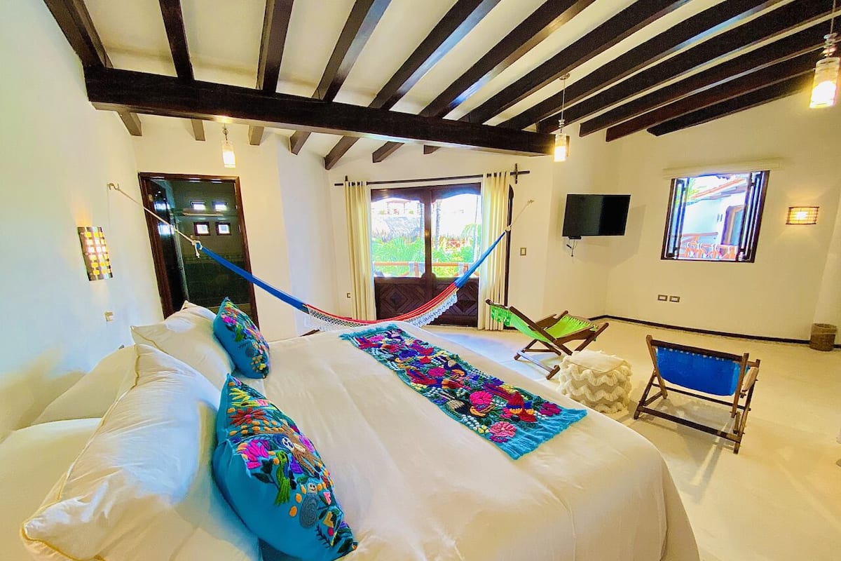 A bedroom with a bed, pillows, a hammock, a television, and a window.