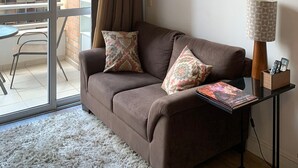 Un salon chaleureux avec un canapé marron, deux coussins à motifs, une table avec une lampe et un tableau représentant un paysage urbain, encadré.