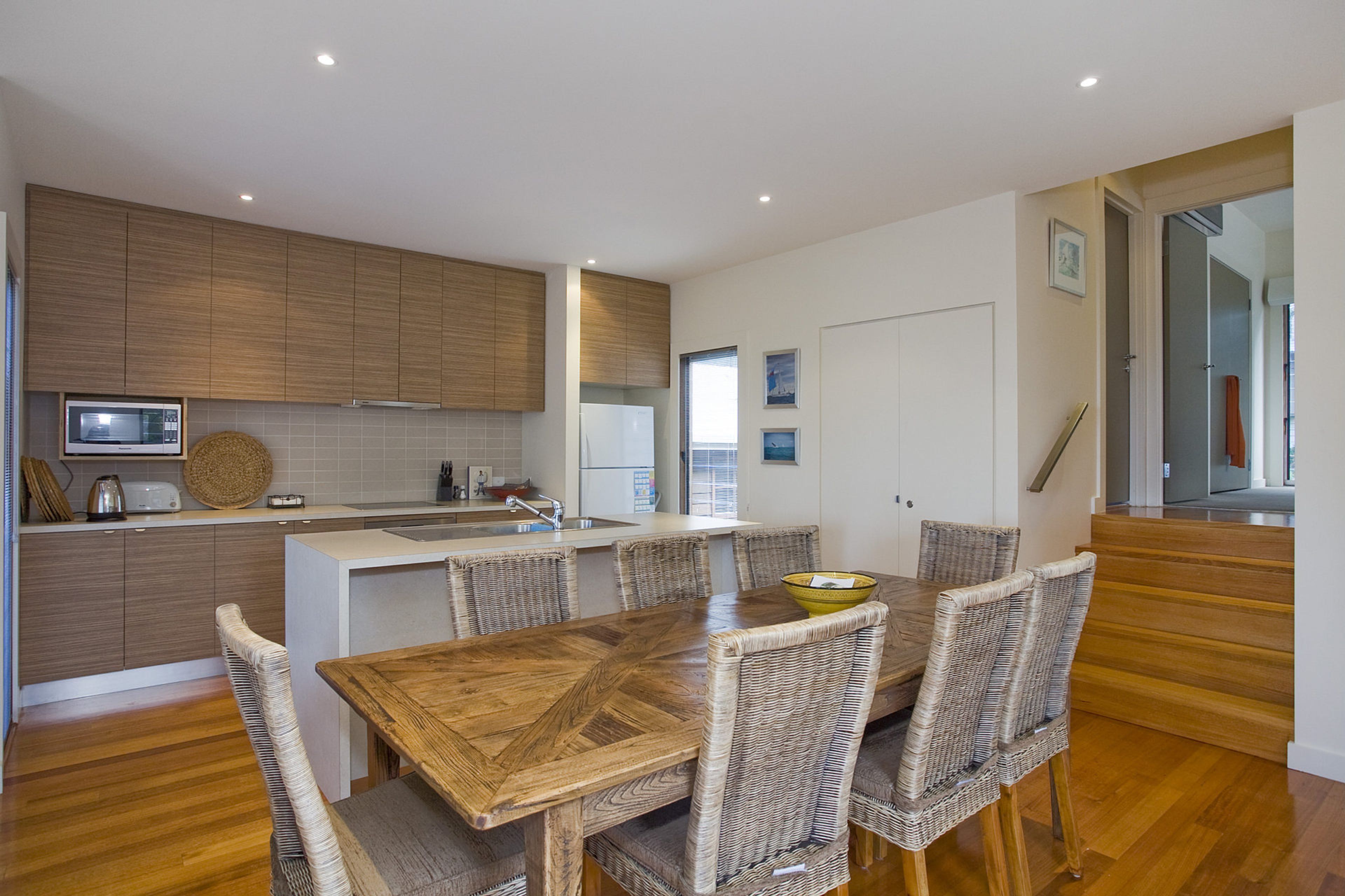 Une cuisine moderne avec des meubles en bois, un four encastré et un coin repas comprenant une table en bois et des chaises en osier.